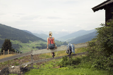 Rückansicht einer Frau, die auf einem Zaun sitzt und auf eine Berglandschaft blickt, Sattelbergalm, Tirol, Österreich - CUF26268