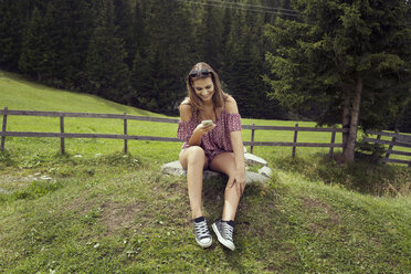 Young woman sitting in field reading smartphone texts, Sattelbergalm, Tyrol, Austria - CUF26269