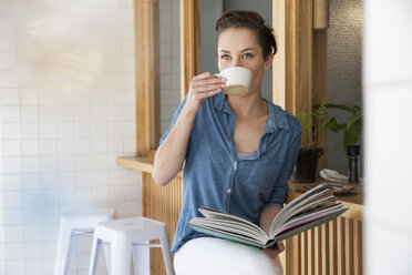 Young woman sitting at bar in cafe, drinking coffee, holding book - CUF26278