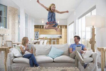 Mädchen springt mitten in der Luft vom Wohnzimmersofa, während die Eltern ein digitales Tablet benutzen - CUF26437