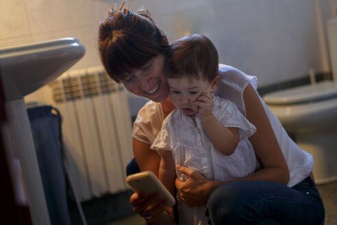 Mittlere erwachsene Frau und kleine Tochter schauen im Badezimmer auf ihr Smartphone - CUF26478