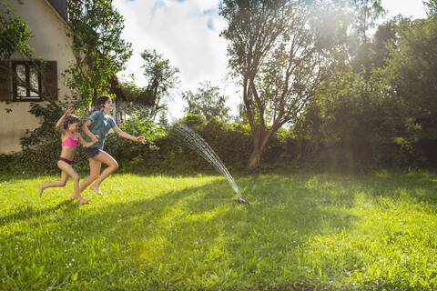 Mädchen und ihre kleine Schwester haben Spaß mit Rasensprenger im Garten, lizenzfreies Stockfoto