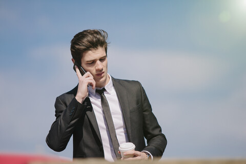 Junger Geschäftsmann plaudert mit seinem Smartphone, während er Kaffee zum Mitnehmen trinkt, lizenzfreies Stockfoto