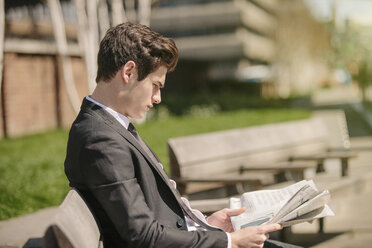 Young city businessman sitting on bench reading newspaper - CUF27114