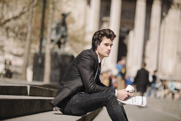 Young city businessman with newspaper sitting on bench listening to headphones - CUF27117