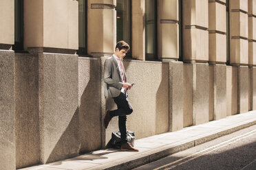 Junger Geschäftsmann lehnt an einem Bürogebäude und liest einen Text auf seinem Smartphone - CUF27119