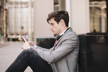 Junger Geschäftsmann sitzt auf dem Bürgersteig und liest Smartphone-Nachrichten - CUF27122