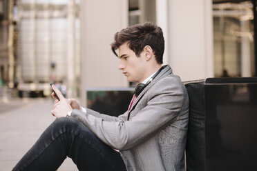 Young city businessman sitting on sidewalk reading smartphone messages - CUF27122