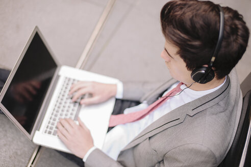 Over shoulder view of young city businessman sitting on sidewalk typing on laptop - CUF27124