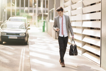 Junger Geschäftsmann in der Stadt liest Texte auf seinem Smartphone, während er vor seinem Büro spazieren geht - CUF27128