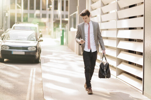 Young city businessman reading smartphone texts whilst walking outside office - CUF27128