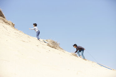 Zwei Jungen klettern auf einen sandigen Hügel und ziehen einen Schlitten hinter sich her - CUF27252