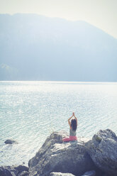 Mid adult woman, sitting on rock, beside lake, in yoga position, rear view - CUF27294