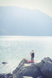 Mittlere erwachsene Frau, auf einem Felsen sitzend, neben einem See, in Yogaposition, Rückansicht - CUF27294