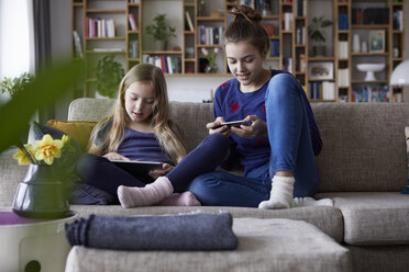 Two sisters sitting on sofa, playing and catting with digital devices - RBF06240
