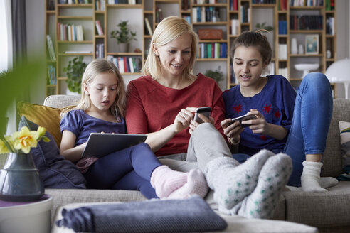 Mother and her daughters sitting on couch, having fun using digital laptop and playing with smartphones - RBF06243