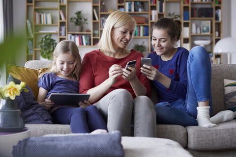 Mutter und ihre Töchter sitzen auf der Couch und haben Spaß an einem digitalen Laptop und spielen mit Smartphones, lizenzfreies Stockfoto