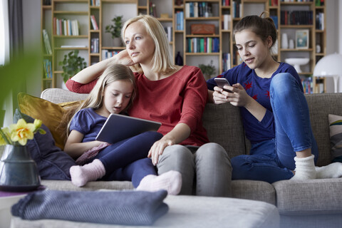 Traurige Mutter sitzt mit ihren Töchtern auf der Couch und spielt mit mobilen Geräten, lizenzfreies Stockfoto