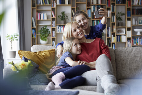 Mutter und ihre Töchter sitzen auf der Couch und machen Smartphone-Selfies, lizenzfreies Stockfoto