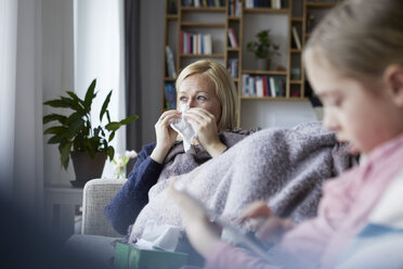 Mother sitting on couch, having a cold, daughter playing in foreground - RBF06276