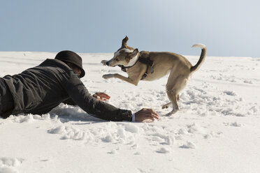 Mann spielt mit Hund im Winter, auf Schnee liegend - REAF00273