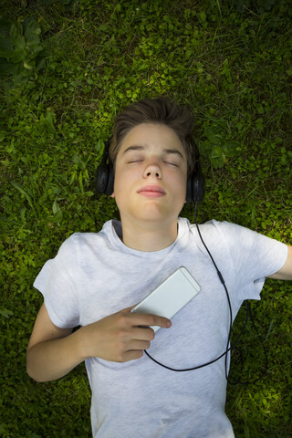 Porträt eines auf der Wiese liegenden Jungen, der mit Kopfhörern und Smartphone Musik hört, lizenzfreies Stockfoto