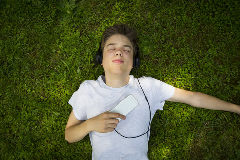 Porträt eines auf der Wiese liegenden Jungen, der mit Kopfhörern und Smartphone Musik hört - LVF07077