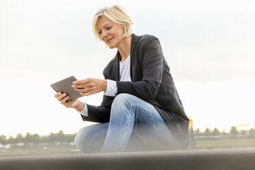 Smiling senior businesswoman sitting with tablet outdoors - FMKF05137