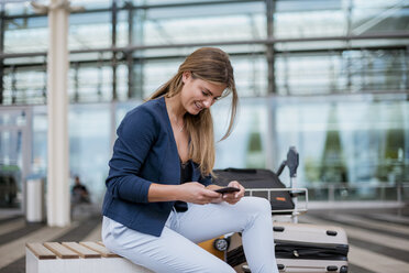 Smiling young businesswoman sitting outdoors with cell phone and suitcase - DIGF04606