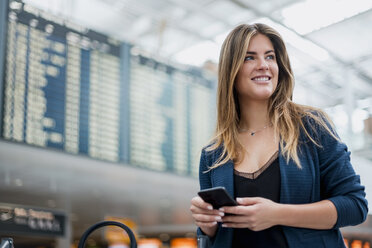 Lächelnde junge Frau mit Handy an der Abfahrtstafel, die sich umschaut - DIGF04613