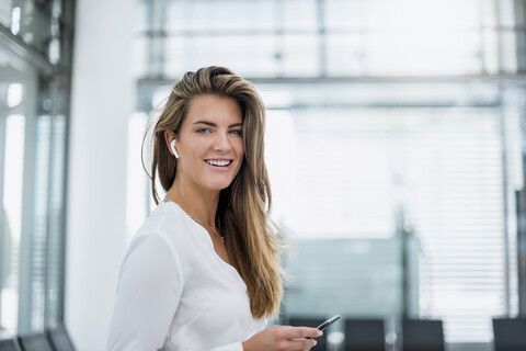 Lächelnde junge Frau mit In-Ohr-Telefon, die ein Mobiltelefon benutzt, lizenzfreies Stockfoto