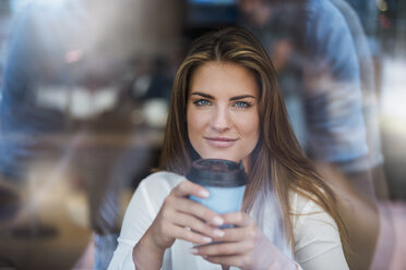 Portrait of smiling young woman behind windowpane with takeaway coffee - DIGF04648