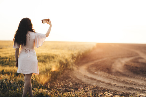 Junge Frau steht am Feld und macht ein Selfie bei Sonnenuntergang, lizenzfreies Stockfoto
