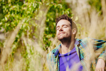 Mid adult man sitting in tall grass eyes closed basking in sunlight - CUF29186