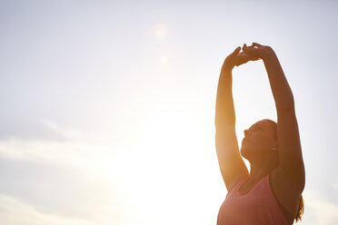 Young female runner stretching arms against sunlit sky - CUF29380