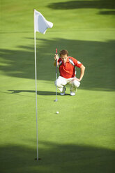 Vorderansicht eines Golfspielers, der sich vor der Golffahne hinhockt und seine Strategie überlegt - CUF29419