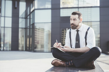 Stilvoller Geschäftsmann sitzt im Schneidersitz und macht Tagebuchnotizen auf seinem Smartphone außerhalb des Büros - CUF29477