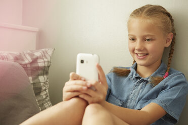 Mädchen sitzt an die Wand gelehnt und schaut lächelnd auf ihr Smartphone - CUF29514