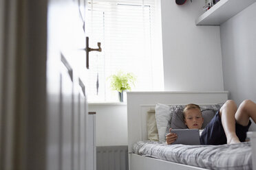 View through doorway of boy lying on bed looking down at digital tablet - CUF29541