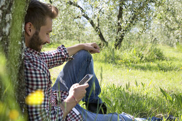 Junger Mann sitzt an einen Baum gelehnt und benutzt ein Smartphone, während er lächelnd nach unten schaut - CUF29652