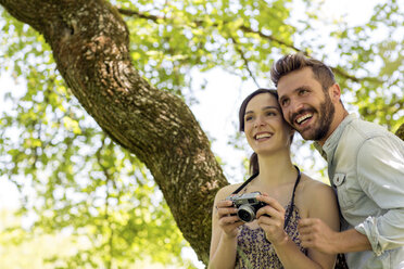 Junges Paar unter einem Baum, das die Kamera hält und lächelnd wegschaut - CUF29671