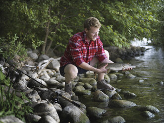 Young fisherman crouching on riverbank holding fish, Copenhagen, Denmark - CUF29983