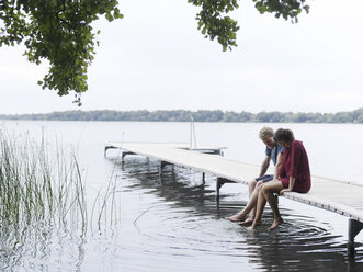 Paar sitzt auf dem Pier nebeneinander und taucht die Zehen ins Wasser, Kopenhagen, Dänemark - CUF29984