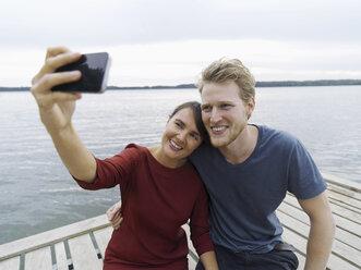 Couple on pier side by side using smartphone to take selfie smiling, Copenhagen, Denmark - CUF29987