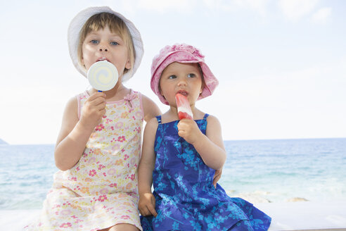 Weibliches Kleinkind und Schwester mit Sonnenhüten essen Eislutscher am Strand - CUF30110