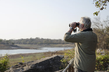 Älterer Mann schaut durch ein Fernglas auf einen Fluss, Kafue National Park, Sambia - CUF30122