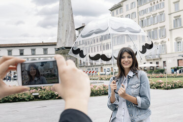 Junge Frau mit Regenschirm posiert für einen Freund, der ein Smartphone zum Fotografieren benutzt, Piazza Santa Maria Novella, Florenz, Toskana, Italien - CUF30153