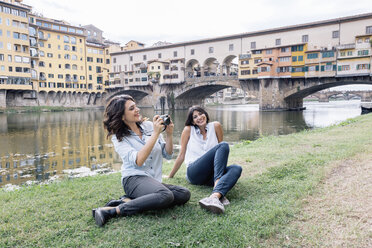 Lesbian couple sitting on arno river bank in front of Ponte Vecchio holding digital camera smiling, Florence, Tuscany, Italy - CUF30169