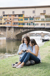 Lesbisches Paar, das zusammensitzt und in die Digitalkamera schaut, vor der Ponte Vecchio lächelnd, Florenz, Toskana, Italien - CUF30170