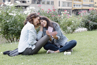 Lesbian couple sitting on grass next to Arno river giving, receiving gift, Florence, Tuscany, Italy - CUF30174